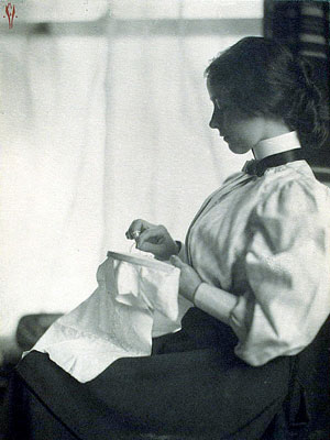 Photograph shows a profile view of a young woman, seated by a window, embroidering fabric held in an embroidery frame.